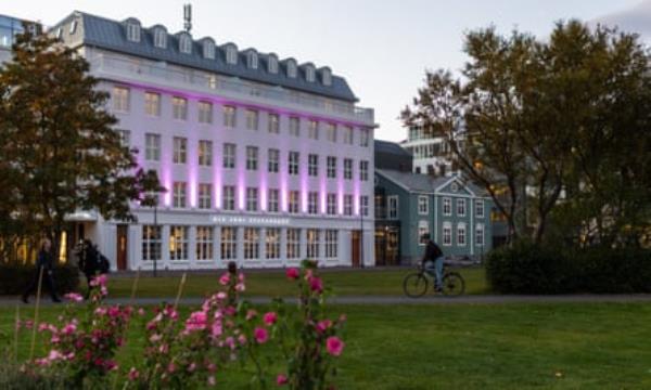 A cyclist rides past a large white building with pink uplighters illuminating it